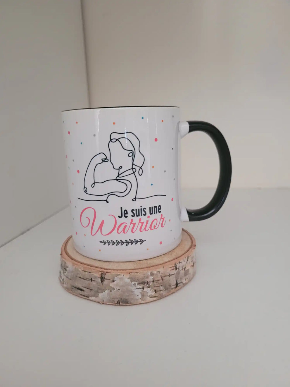 Tasse en céramique blanche avec une poignée et un bord noirs, présentant un motif en dessin au trait d'un bras musclé et les mots ’Je suis une Guerrière’ en rose.
