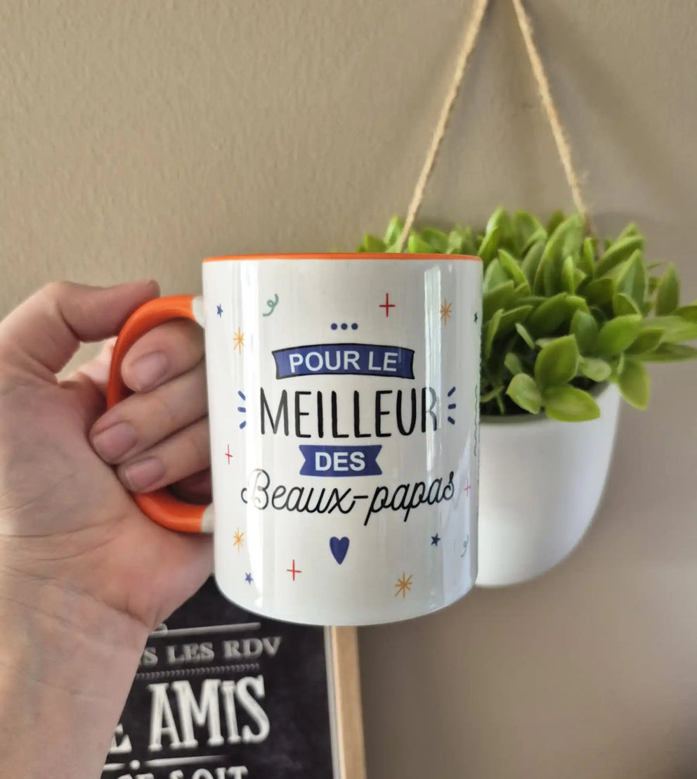 Une tasse en céramique blanche avec une poignée et un rebord orange, décorée d'accents bleus et orange et la phrase ’POUR LE MEILLEUR DES Beaux-papas’ en français.